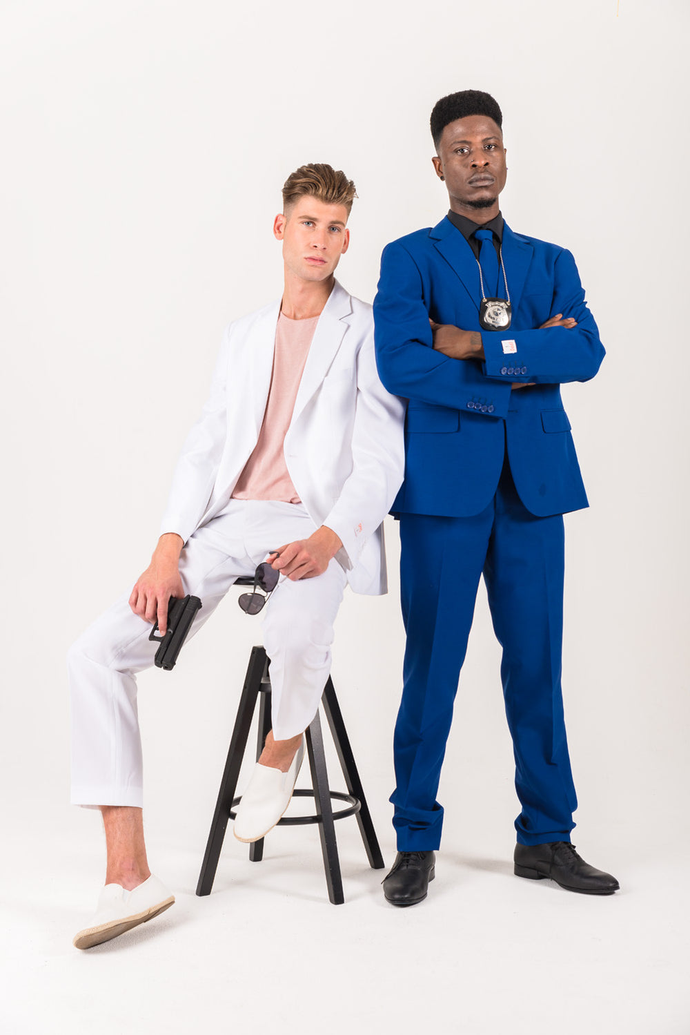 Two men in suits, one seated in white holding sunglasses and a handgun, and one standing in blue with his arms crossed and a badge necklace.