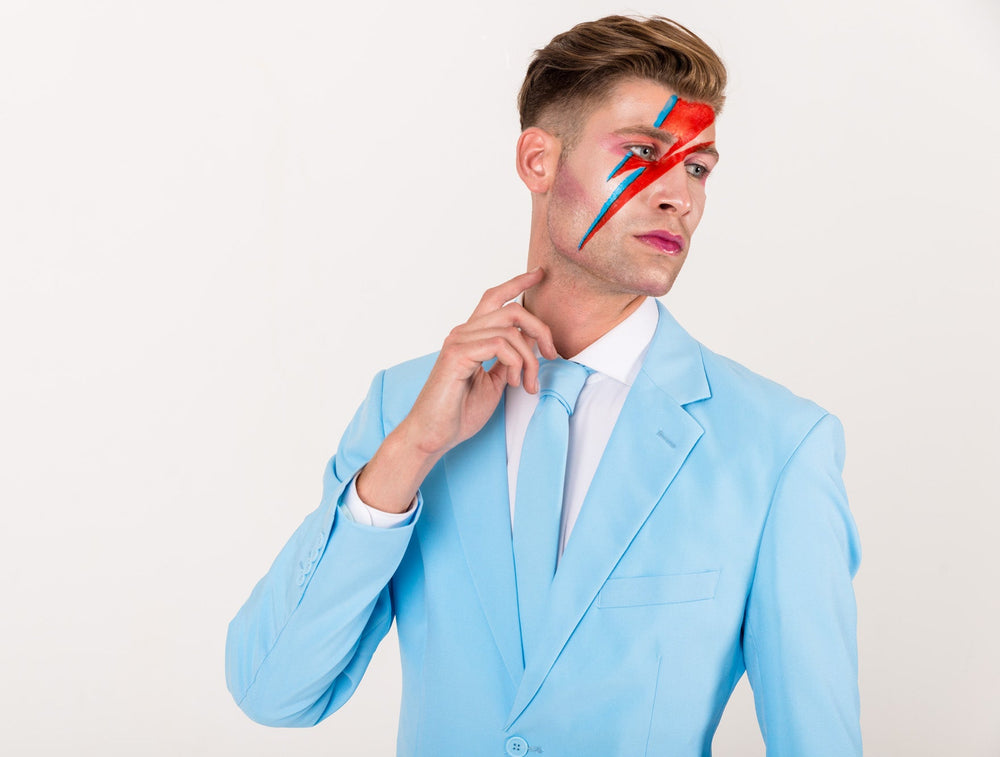 A man wearing a light blue suit, white dress shirt, and matching tie, with red and blue lightning-bolt face paint on his forehead, posing against a plain white background.