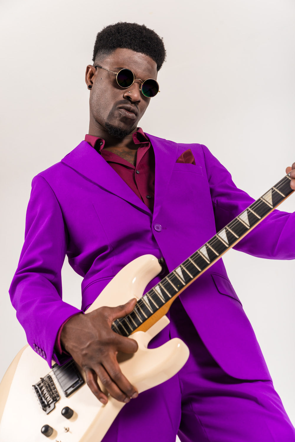 Man in a bright purple suit and sunglasses playing a white electric guitar.