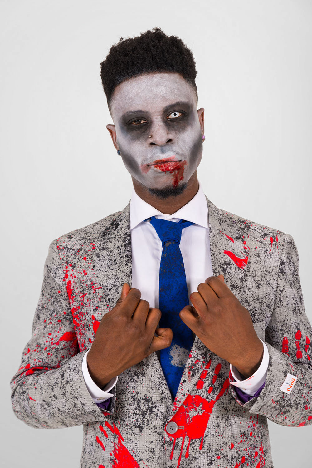 Man in a gray suit with red blood splatter, blue tie, and zombie makeup.