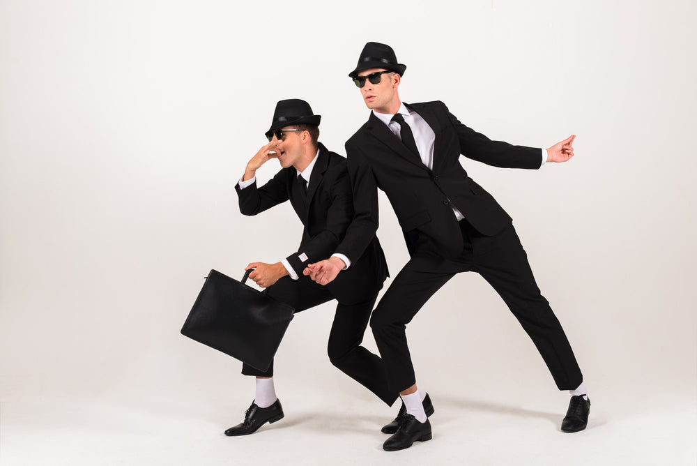 Two men in black suits, hats, and sunglasses posing playfully, with one carrying a black briefcase. They imitating he blues brothers