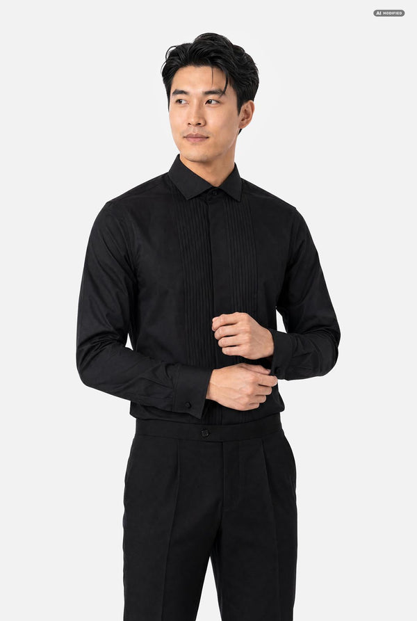 A man wearing a black long-sleeve pleated tuxedo shirt and matching black trousers, standing against a light gray studio background.