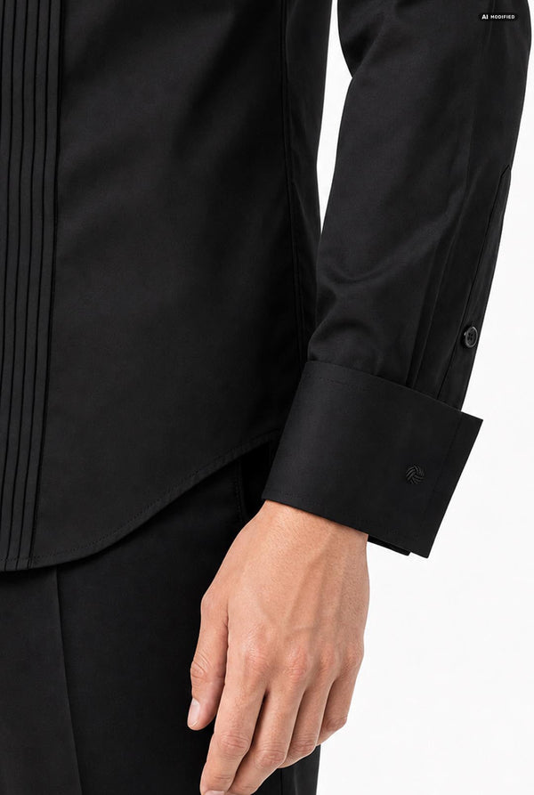 Close-up of a person wearing a black formal tuxedo shirt with pleated front panels and a buttoned cuff, paired with black trousers, against a clean grey background.