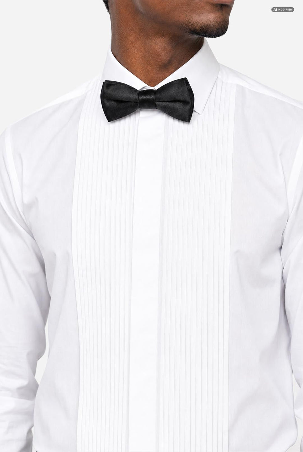 A man wearing a white long-sleeve tuxedo shirt with black bow tie and black trousers, adjusting his cuffs against a light gray studio background.