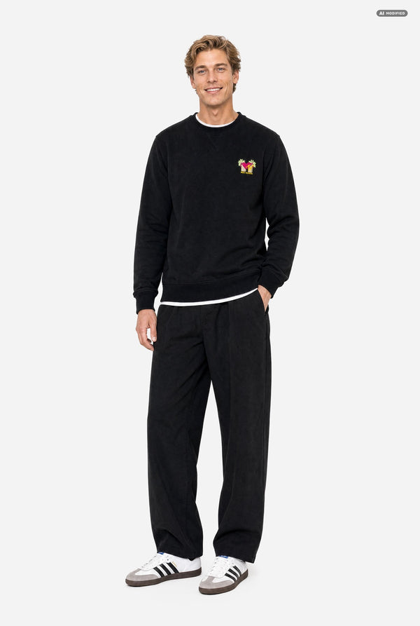 A man wearing a black crewneck sweatshirt with a small colorful embroidered MTV graphic on the left chest, paired with black sweatpants, against a light gray background. Full body image.