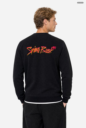 A man wearing a black crewneck sweatshirt with a colorful embroidered Spring Break MTV graphic on the back, paired with black sweatpants, against a light gray background.