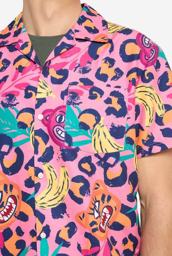 A man wearing a vibrant two-piece summer outfit featuring a bold tropical print. The short-sleeve button-up shirt and matching shorts showcase a lively design with neon pink, orange, and yellow leopard prints, bananas, green palm leaves, and quirky cartoon faces. The outfit is styled casually with a green T-shirt underneath and is perfect for summer parties, vacations, or poolside fun. Chest close up.