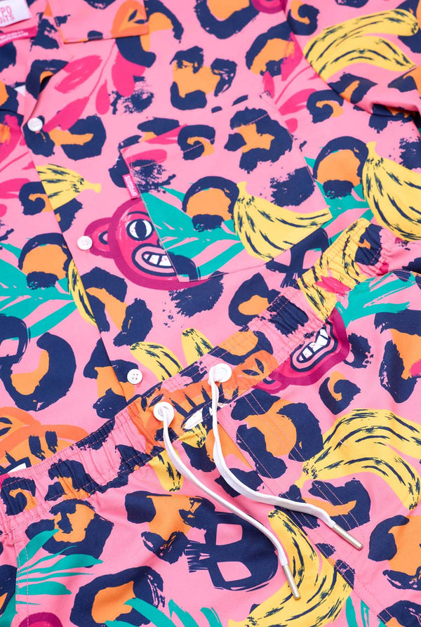 Image of a vibrant two-piece summer outfit featuring a bold tropical print. The short-sleeve button-up shirt and matching shorts showcase a lively design with neon pink, orange, and yellow leopard prints, bananas, green palm leaves, and quirky cartoon faces. The outfit is styled casually with a green T-shirt underneath and is perfect for summer parties, vacations, or poolside fun. Close-up.