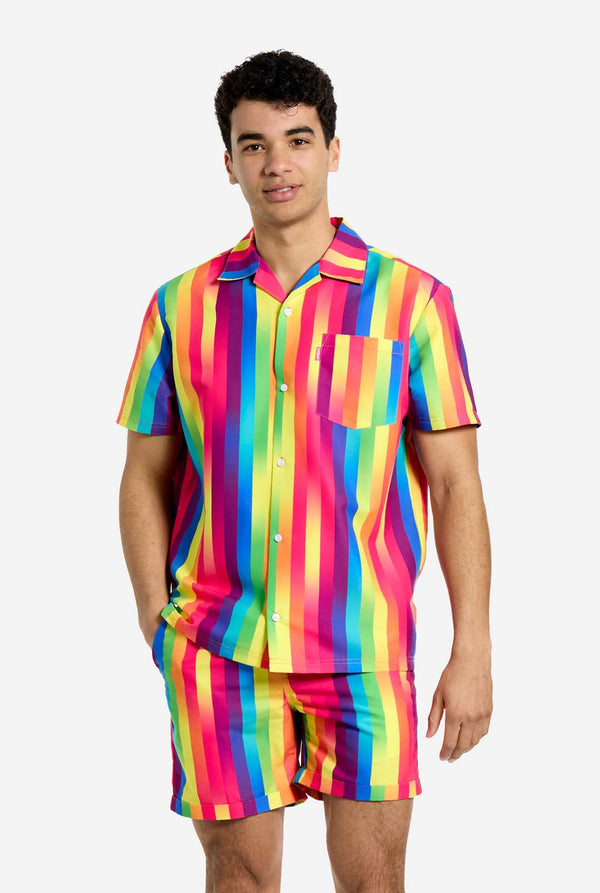 A man is wearing a vertically striped rainbow-colored summer set, consisting of shirt and shorts. He is smiling in the camera and holding one hand in his pocket.