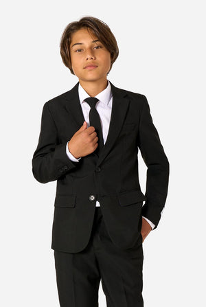 Teenager wearing a black suit and a white shirt on a light background.