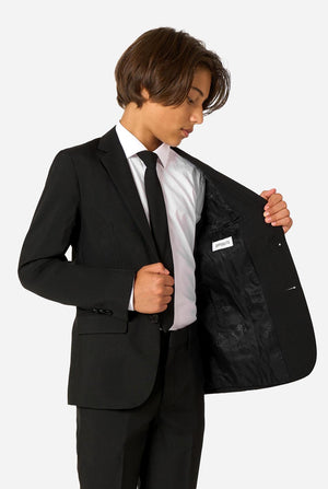 Teenager wearing a black suit and a white shirt on a light background. Holding one side of the jacket open.