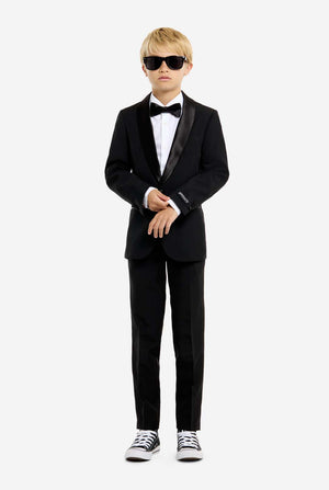 Person wearing a black tuxedo with sunglasses and white sneakers on a white background