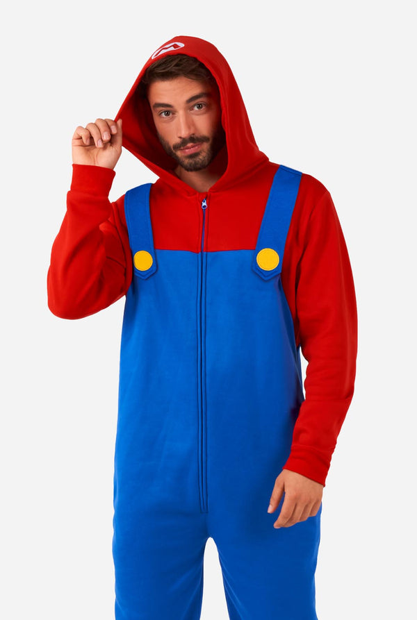 A man is dressed in a Mario-themed onesie. The outfit look as a bright red hoodie with an "M" symbol on the hood and blue overalls with large yellow buttons on the straps. He is holding one side of the hood, as if pulling it over his head, and looking directly at the camera with a neutral expression. The overall design mimics the look of the popular video game character Mario, with contrasting red and blue colors. The background is plain, putting focus on the costume and the man's playful pose.