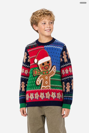 A young boy wearing a festive Christmas knit sweater with a gingerbread man design, paired with khaki pants, photographed against a solid light gray background.