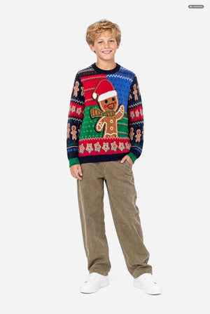 A young boy modeling a festive Christmas sweater with a gingerbread man design, paired with brown corduroy pants and white sneakers, set against a plain light gray background.