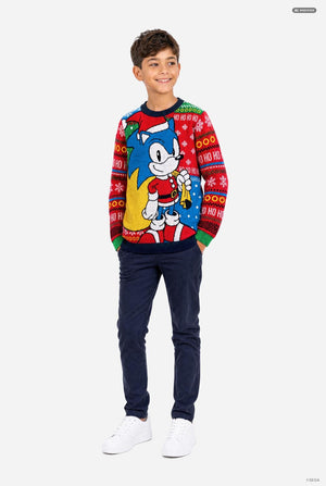 A young boy modeling a festive red Christmas sweater featuring Sonic the Hedgehog, paired with navy blue pants and white sneakers, set against a plain light gray background.