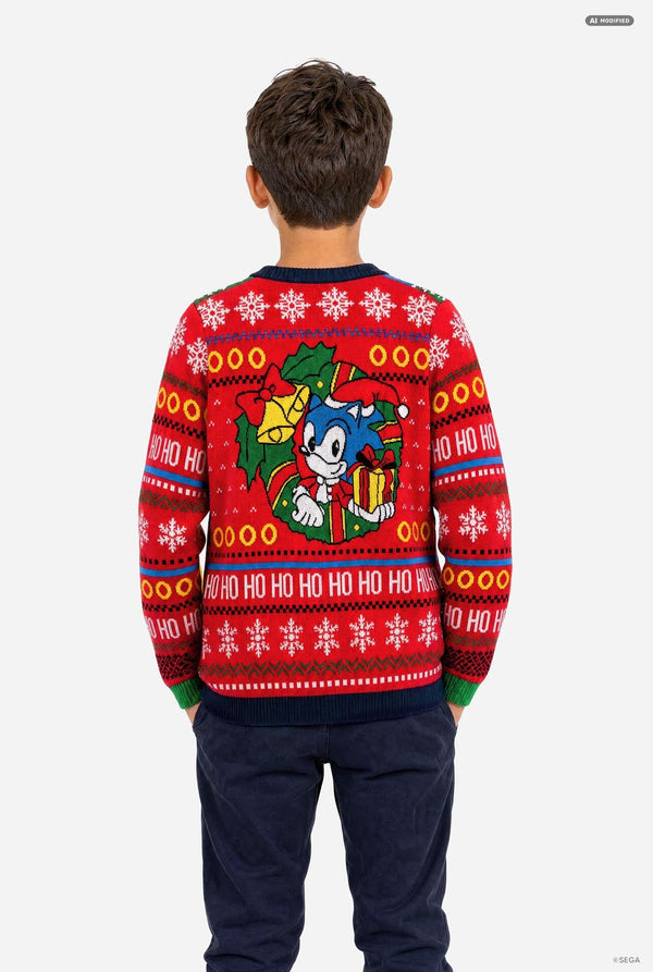 A child viewed from the back wearing a red Christmas sweater featuring Sonic the Hedgehog graphics, including a large Sonic character print and 'HO HO HO' patterns, paired with navy pants against a light gray background.