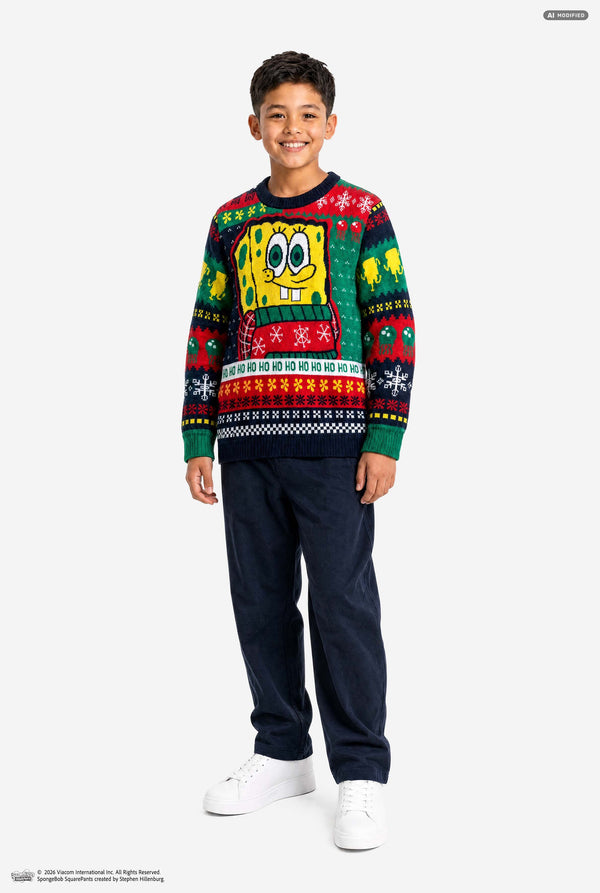 A young boy modeling a long-sleeve crewneck holiday sweater featuring SpongeBob SquarePants, paired with dark navy pants and white sneakers, against a light gray studio background.