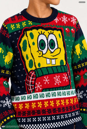 A close-up of a person wearing a colorful SpongeBob-themed Christmas sweater. The knit design features a large yellow SpongeBob character with green eyes and a red patterned scarf, surrounded by holiday motifs including snowflakes, bells, and the phrase 'HO HO HO' across the lower front.