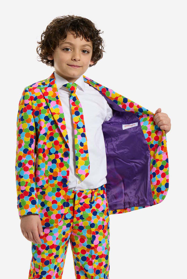 In this image, a kid is wearing a boys’ suit with all-over multicolor confetti print and white shirt. The suit has a bright pattern with circles in various colors including orange, yellow, pink, green, blue, and red. Kid holding one side of the jacket open, showing purple inside. 