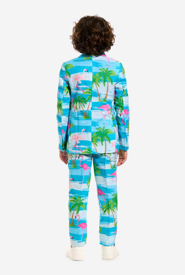 In this image, a kid is wearing a light blue boys’ suit with a tropical print and a white shirt. The suit has a blue print resembling water and pink flamingos and green palm trees. View from the back.