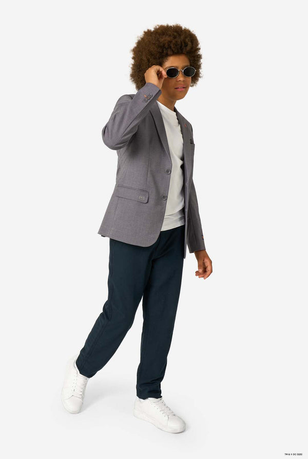 A child wearing a grey blazer and sunglasses, walking confidently in dark trousers and white sneakers.