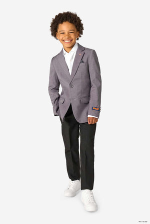 A young boy wearing a grey blazer and black trousers, smiling and standing with his hands in his pockets.