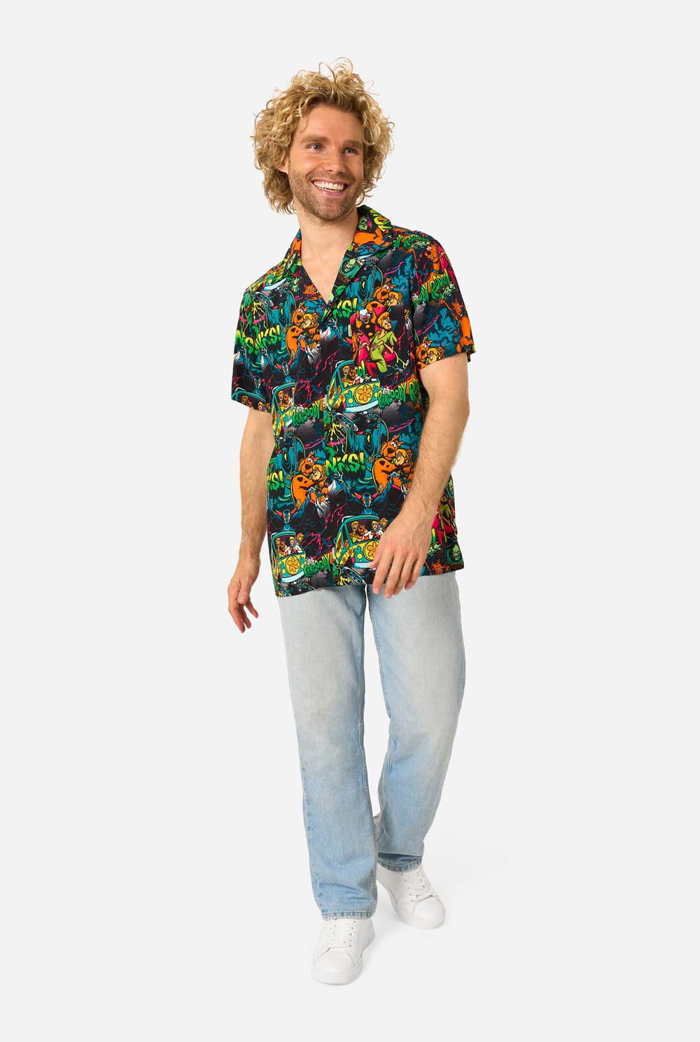 A man with curly blonde hair wearing a short-sleeve Hawaiian shirt with a vibrant, colorful, graphic Scooby-Doo print on a black background, paired with light blue jeans, against a plain light gray background.