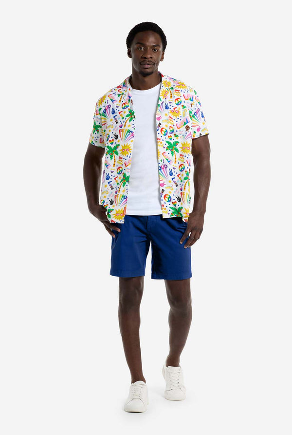A man is wearing a white Hawaiian shirt with a rainbow pride print, Print consists of peace symbols, hearts and other icons, Also, wearing blue shorts. He is looking in the camera.