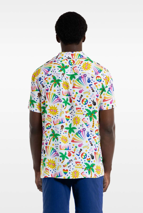 A man is wearing a white Hawaiian shirt with a rainbow pride print, Print consists of peace symbols, hearts and other icons, Also, wearing blue pants. He is facing away from the camera and showing the back side of the shirt.