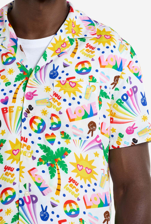 A man is wearing a white Hawaiian shirt with a rainbow pride print, Print consists of peace symbols, hearts and other icons. Chest close up.