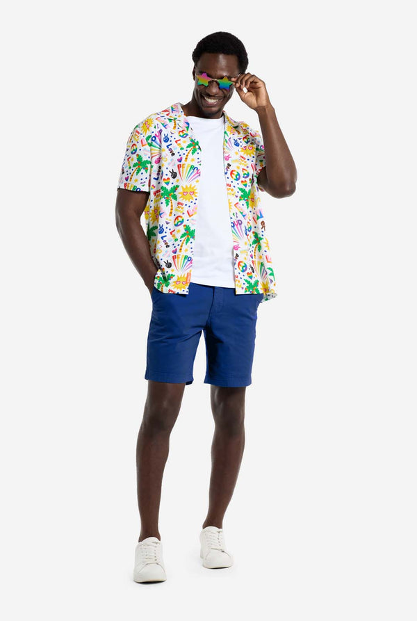 A man is wearing a white Hawaiian shirt with a rainbow pride print, Print consists of peace symbols, hearts and other icons. Also, wearing blue shorts. He is smiling in the camera and wearing star shaped rainbow sunglasses.