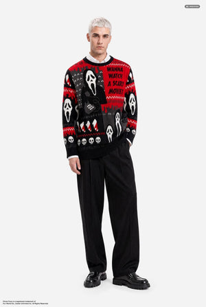 A male model with platinum blonde hair wearing a black and red Halloween-themed knit sweater with Ghostface and skull patterns, paired with black pants, against a light gray background. Full body image.