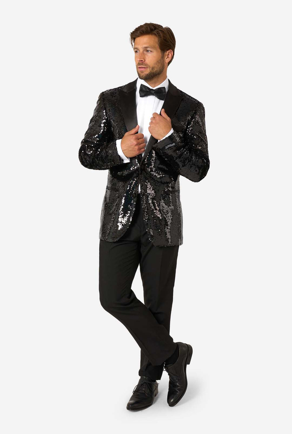 A man modeling a black sequined tuxedo jacket with black satin lapels, worn over a white dress shirt and black bow tie, against a light gray studio background. A circular inset at the lower left shows a close-up of the black sequin texture.