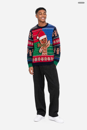 A man modeling a festive Christmas sweater with a gingerbread man design, paired with black trousers and white sneakers, against a light gray background.