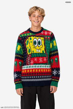 A young man wearing a SpongeBob-themed Christmas sweater, standing against a plain light gray studio background. The sweater features a large SpongeBob graphic on the chest and festive multicolor holiday patterns including snowflakes, bells, and 'HO HO HO' text.