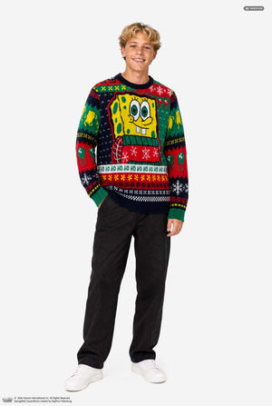 A young man wearing a SpongeBob-themed Christmas sweater, standing against a plain light gray studio background. The sweater features a large SpongeBob graphic on the chest and festive multicolor holiday patterns including snowflakes, bells, and 'HO HO HO' text. Full body image.