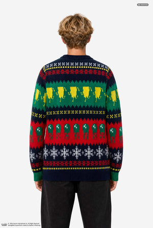 A young man wearinga  festive Christmas sweater against a light gray background. The back of the long-sleeve knit sweater features a multi-colored holiday pattern with rows of yellow lemons, green olives, red peppers, and white snowflakes on a dark navy base.