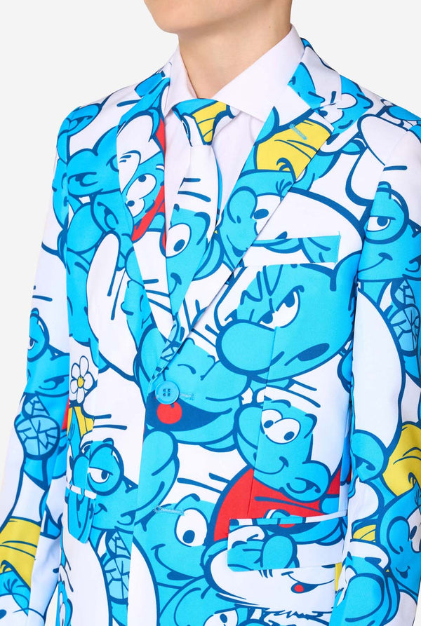 Teen boy wearing a vibrant Smurfs kids suit with colorful cartoon character prints in blue, red, yellow, and white, paired with a matching tie and white shirt, smiling against a plain background. Chest close up.
