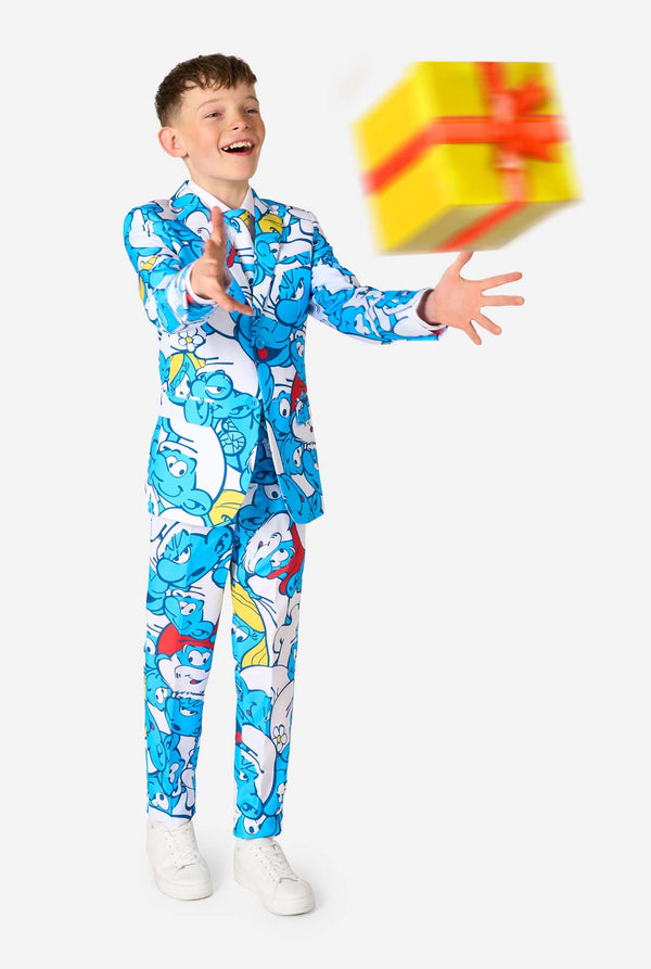 Teen boy wearing a vibrant Smurfs kids suit with colorful cartoon character prints in blue, red, yellow, and white, paired with a matching tie and white shirt, smiling against a plain background. Throwing a present.
