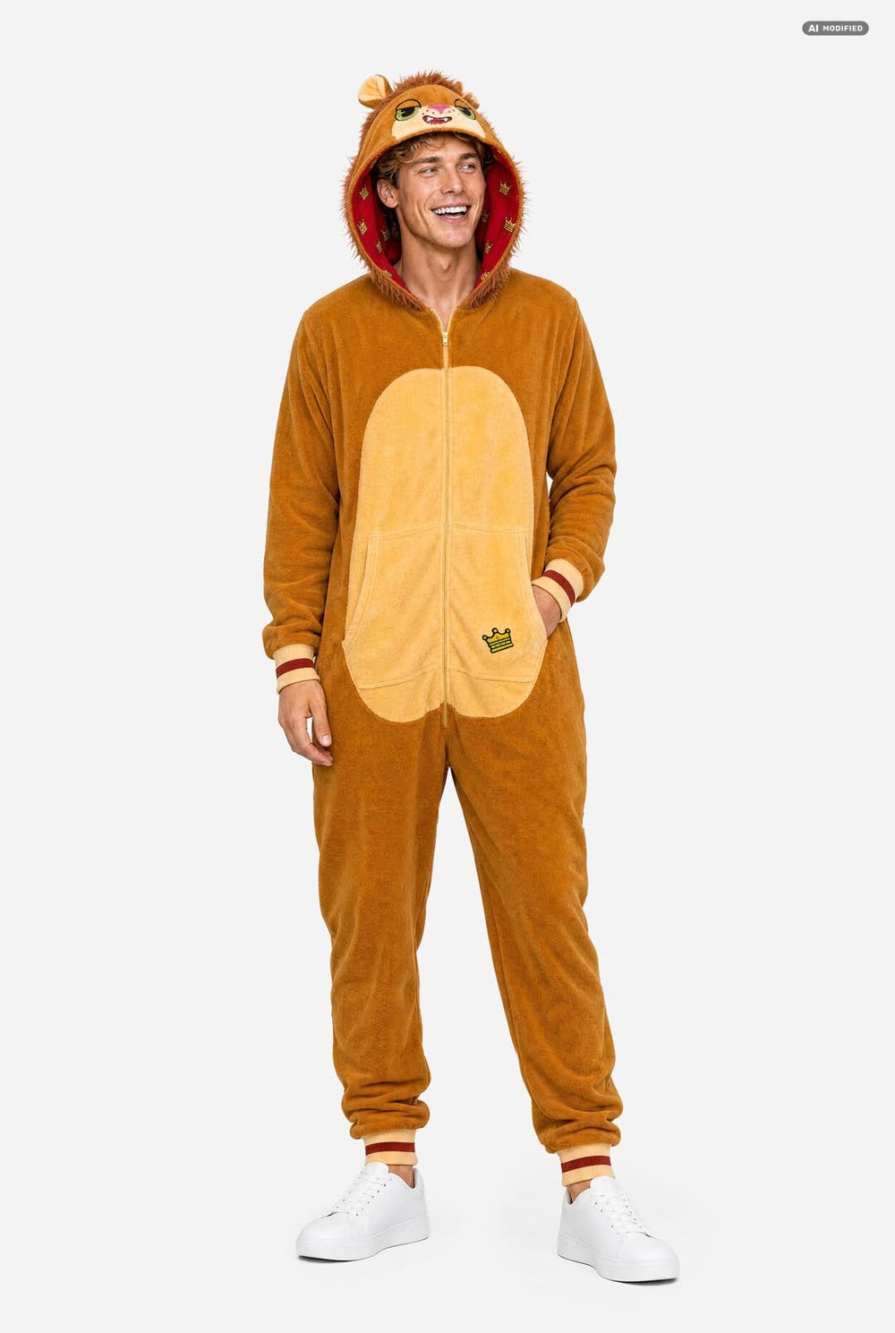 A man wearing a plush lion onesie onesie costume with a hood featuring a lion face and ears, a tan body, and a cream-colored belly patch, photographed against a light gray studio background.