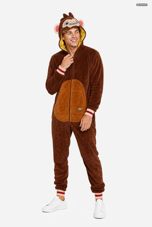 A man modeling a brown plush animal onesie with a hood featuring a monkey face, pink ears, and a yellow inner lining. The one-piece has a front zipper, a tan patch on the chest, and red-and-white striped cuffs, shown against a light gray background.