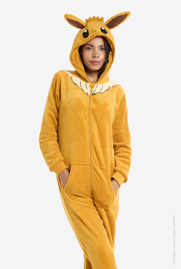 A woman is wearing an adorable Eevee onesie, modeled after the beloved Pokémon character. This adult unisex onesie is designed in a warm brown color, resembling Eevee’s fur, and features a fluffy beige collar mimicking Eevee’s mane. The hood includes Eevee's signature large brown ears, big black eyes. The onesie has a front zipper closure and convenient pockets at the front, offering both comfort and ease of wear. Hands in pockets.