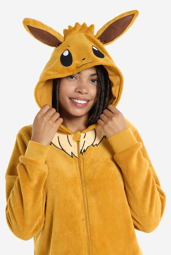 A woman is wearing an adorable Eevee onesie, modeled after the beloved Pokémon character. This adult unisex onesie is designed in a warm brown color, resembling Eevee’s fur, and features a fluffy beige collar mimicking Eevee’s mane. The hood includes Eevee's signature large brown ears, big black eyes. The onesie has a front zipper closure and convenient pockets at the front, offering both comfort and ease of wear. View of the upper part and holding the hood with two hands.