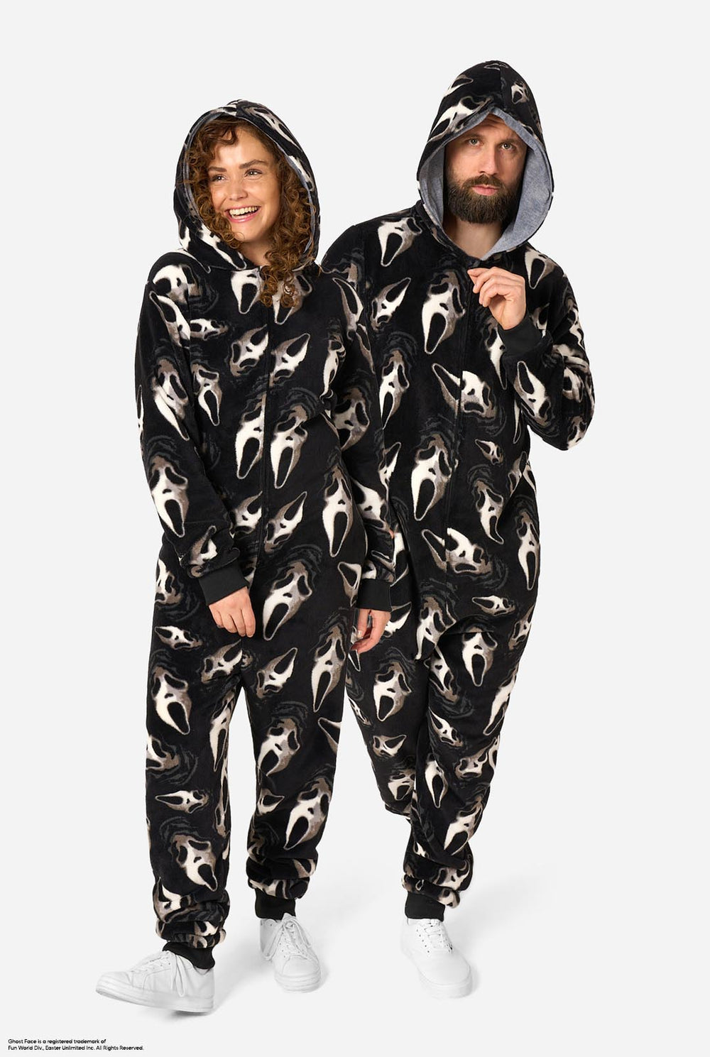 A man and a woman modeling matching black hooded onesie pajamas with an all-over white Ghost Face print, standing against a light gray studio background.
