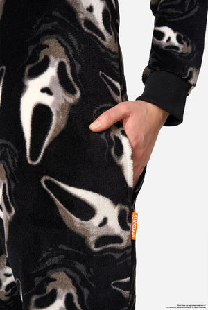 Close-up of a person wearing a black fleece onesie or jumpsuit with a Ghostface-style mask print in white and gray, showing a hand in a side pocket against a light gray background.