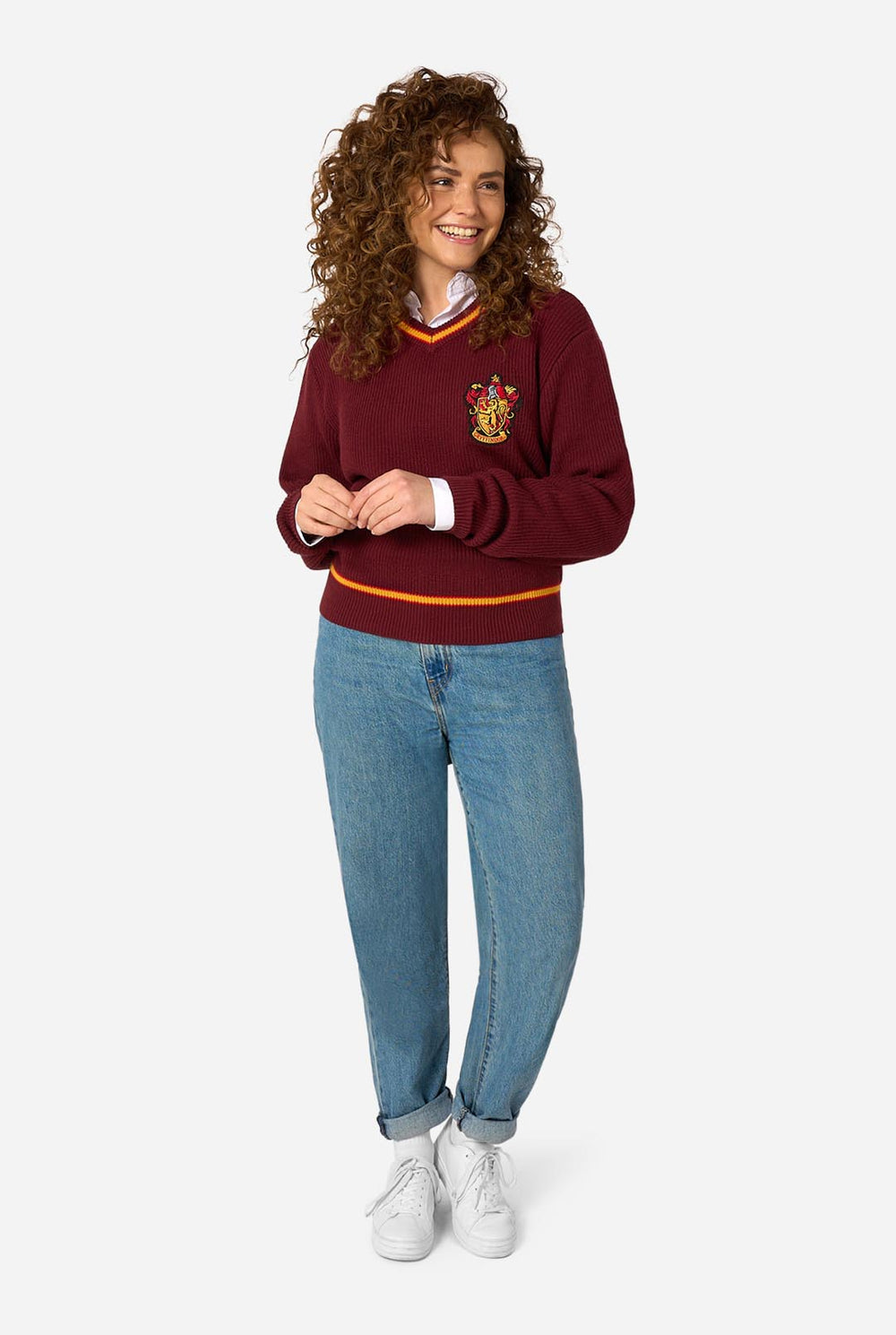 A man wearing a maroon ribbed V-neck Harry Potter Gryffindor sweater with yellow and orange trim, featuring the Gryffindor house crest on the left chest, styled over a white collared shirt and dark trousers, set against a plain light gray background.
