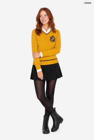 A woman modeling a mustard yellow long-sleeve sweater with a black and white V-neck collar, a black crest patch on the left chest, a black pleated mini skirt, black tights, and black lace-up boots, set against a plain light gray background.