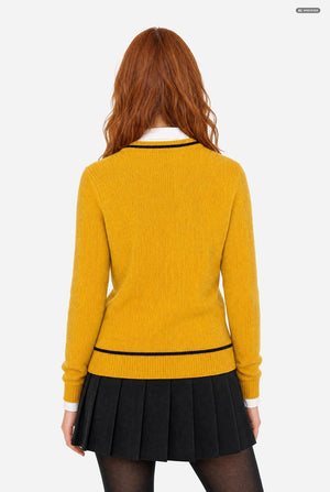 A woman with red hair is shown from the back wearing a mustard yellow ribbed knit sweater with black trim, a black pleated skirt, and black tights against a light gray background.