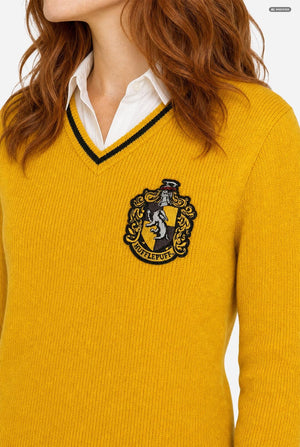 A close-up of a woman wearing a mustard yellow ribbed knit sweater with a black and white V-neck trim and an embroidered Hogwarts crest on the chest, layered over a white collared shirt.